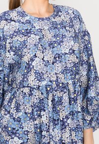 Žena v modrých květovaných šatech s světle modrými, bílými a žlutými květy a sezný zataženým pasem.