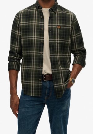 Mann trägt ein dunkelgrünes und graues kariertes Hemd über einem beigen T-Shirt, kombiniert mit blauen Jeans, braunem Gürtel und goldener Armbanduhr.