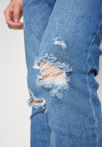 Jeans de mezclilla azul claro con áreas desgastadas en las rodillas y bordes deshilachados. La tela tiene un acabado texturizado con detalles de costura visibles.