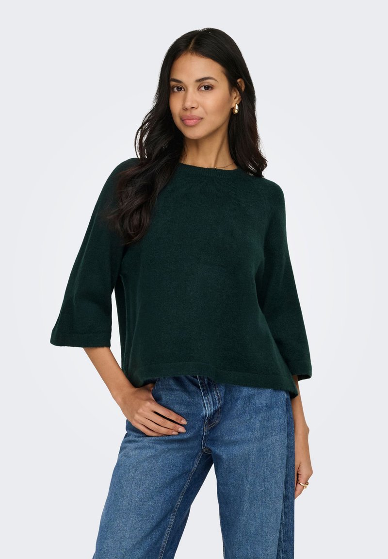Femme aux longs cheveux foncés portant un pull vert foncé et un jean bleu, posant avec une main sur la hanche devant un fond uni.