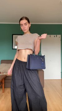 Top cropped riscado em castanho claro e branco, calças largas pretas, segurando uma mala azul-marinho com um charme, em fundo de parede verde.