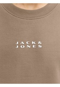 Sudadera marrón con un cuello redondo acanalado. Presenta el logo "JACK & JONES" en letras blancas centradas en la parte frontal. Tejido suave y texturizado.