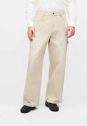 Beige, weit geschnittene Hosen, getragen mit schwarzen Schuhen und einem weißen, langärmeligen Hemd, Hände ruhen an den Seiten.