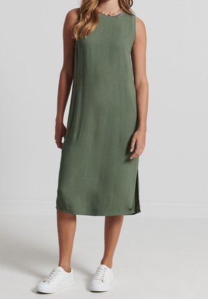 Vestido midi sin mangas de color verde oliva con abertura lateral, combinado con zapatillas blancas y un collar de cadena dorada.