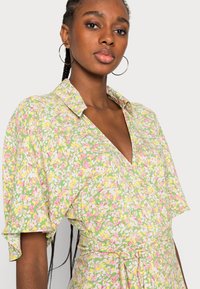 Vestido em formato envelope com fundo verde, apresentando pequenas flores rosas e amarelas. Tem gola, mangas curtas e um detalhe de cintura ajustada com laço.