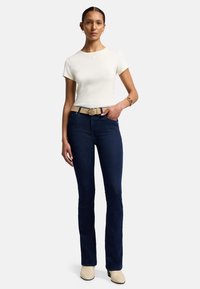 T-shirt bianco a maniche corte abbinato a jeans svasati blu scuro e una cintura beige. La modella indossa stivaletti beige sopra la caviglia, mostrando uno stile semplice e aderente.