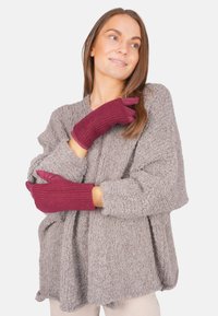 Femme portant un pull gris texturé et des gants en tricot bordeaux, souriant et regardant légèrement sur le côté contre un fond blanc uni.