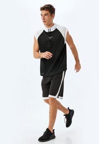 Canotta sportiva nera senza maniche con dettagli bianchi, abbinata a pantaloni corti neri con strisce laterali bianche e scarpe da ginnastica nere con texture.