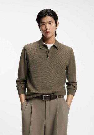 Olive green knit polo shirt with a textured pattern, buttoned collar, and long sleeves, paired with beige trousers and a brown belt.