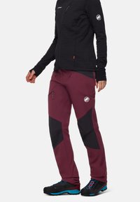 Pantaloni softshell bordeaux con accenti neri, dotati di tasche con zip e cintura elasticizzata, abbinati a una giacca nera con texture e scarpe colorate.
