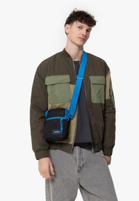 Grøn og khaki bomberjakke med to brystlommer, parret med en lille sort crossbody taske med en blå rem og snørelukning.