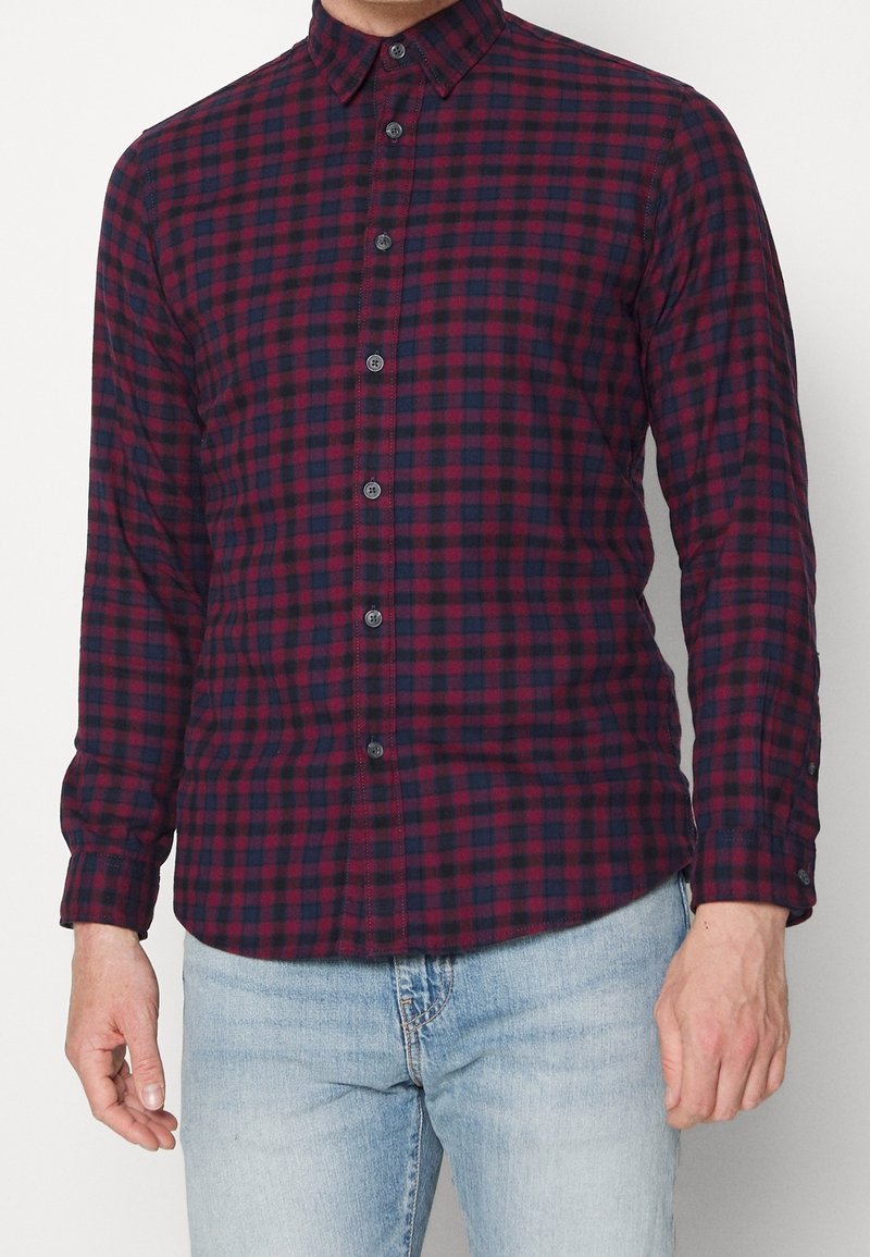 Uomo che indossa una camicia a maniche lunghe aderente con un motivo a scacchi rosso e blu navy e jeans azzurri su uno sfondo neutro.