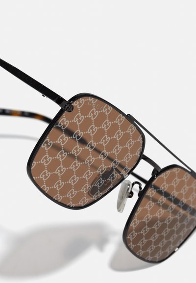 Gafas de sol marrones con un marco de metal negro y lentes estampados que muestran el logo de Gucci. Las formas son angulares con varillas delgadas y delicadas.