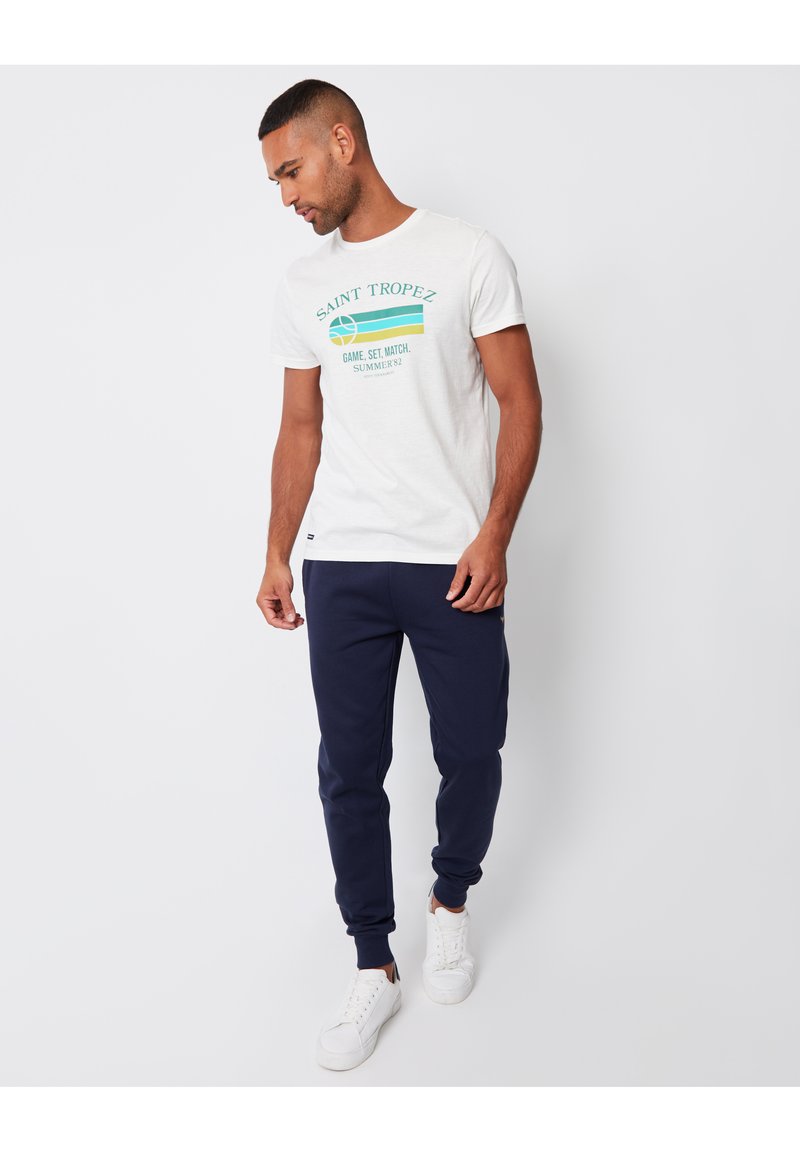 Witte katoen t-shirt met turquoise en gele grafische tekst. Gecombineerd met marineblauwe joggingsbroeken en witte sneakers, met ribgebreide manchetten.