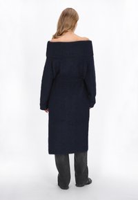 Hosszú, tengerészkék, lecsúszott vállú kötött pulóverruha bordázott textúrával, övvel kiemelt derékkal és egyenes siluettel, laza sötét nadrággal párosítva.
