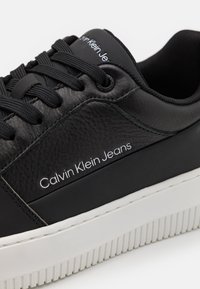 Sneaker nera in pelle con pannello testurizzato, suola in gomma bianca e lacci neri. Presenta il marchio "Calvin Klein Jeans" in bianco.