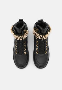 Svarta hög-top sneakers med guldfärgade öglor och dekorativa rhinestones som stavar "MOSCHINO" på varje snörrem.