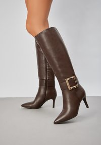 Bottes brunes jusqu'au genou en cuir lisse, avec un bout pointu, un talon aiguille et un grand accroc en métal au niveau de la cheville.