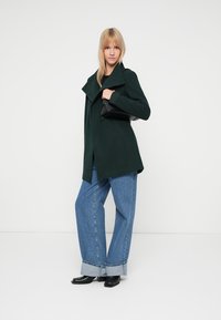 Donkergroene wollen gelaagd jas met een gestructureerde kraag, gecombineerd met losse lichtblauwe jeans en zwarte enkellaarsjes, terwijl ze een zwarte handtas vasthoudt.