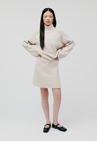 Beiger gerippter Strickpullover mit hohem Kragen, kombiniert mit einem passenden knielangen Rock. Getragen mit schwarzen Ballerinas mit einer Schleife.