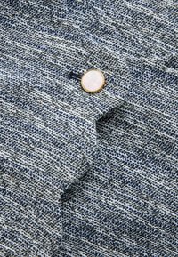 Tessuto blu e bianco con texture e un motivo sottile, caratterizzato da un bottone rotondo con accenti dorati sul bordo.