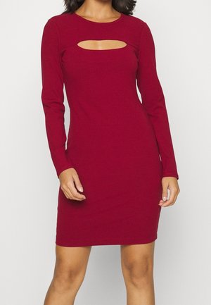 Vrouw draagt een nauwsluitende rode geribde gebreide jurk met lange mouwen en een horizontale uitsparing boven de buste.