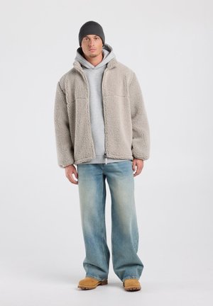 Man met een grijze muts, lichtgrijze hoodie, beige sherpakatoenjas, losse blauwe jeans en beige laarzen, staand tegen een effen lichte achtergrond.