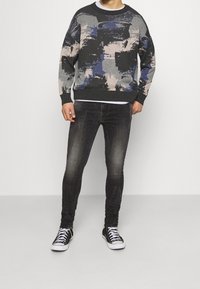 Suéter con un patrón abstracto en negro, azul y beige, material de punto, cuello redondo; combinado con jeans ajustados oscuros y zapatillas negras.