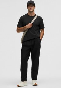 Camiseta negra de manga corta y pantalones negros. Bolsa cruzada beige y zapatillas ligeras con suelas texturizadas. El modelo está de pie contra un fondo gris.
