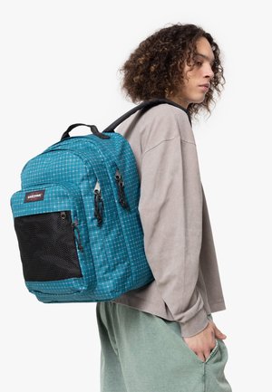 Mochila verde agua con un patrón de cuadrícula, múltiples compartimentos con cremallera y acentos de malla negra. Llevada sobre un suéter gris y pantalones verdes.