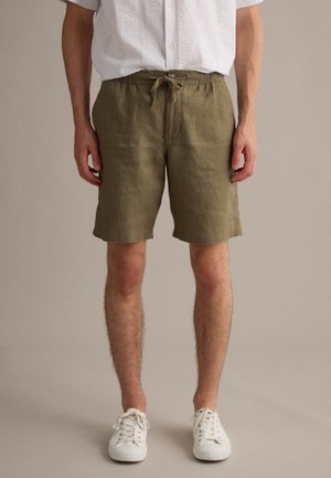 Man draagt olijfgroene short met trekkoord, wit gestructureerd kort shirt en witte sneakers, staand tegen een effen achtergrond.