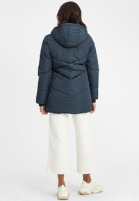 Mørkeblå isolert parka med hette, med quiltet design og elastiske mansjetter, kombinert med hvite cropped bukser og lyse sneakers.