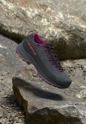 Zapato de senderismo gris con acentos rosas, parte superior texturizada y suela resistente. Presenta una etiqueta GORE-TEX y un collar acolchado para mayor comodidad.