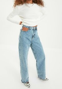 Jeans larges bleu clair avec ourlets effilochés et poches découpées, associés à un haut à manches longues blanc côtelé et des chaussures pointues noir et blanc.