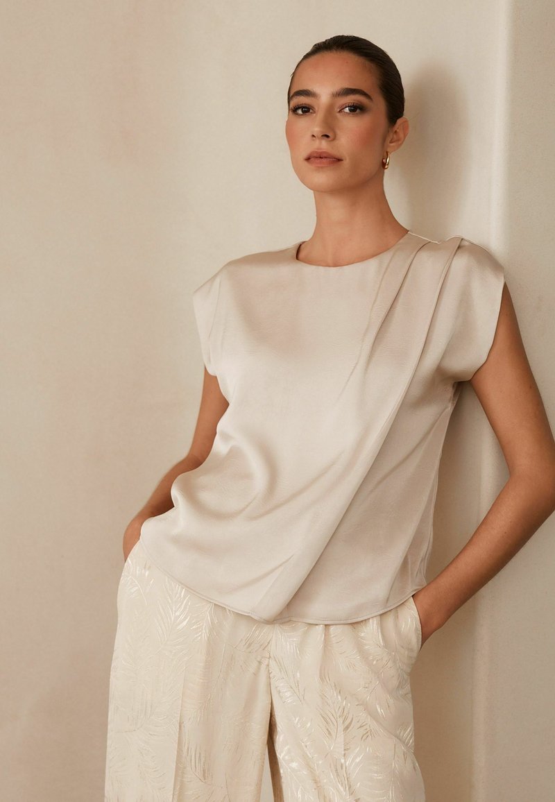 Top de satén beige con un diseño drapeado y mangas con forma de capa, combinado con pantalones de pierna ancha de color claro y brillante que presentan un patrón de hojas.