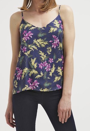 Femme portant un caraco bleu marine avec un imprimé floral jaune et rose et des bretelles fines, associé à un pantalon slim foncé.