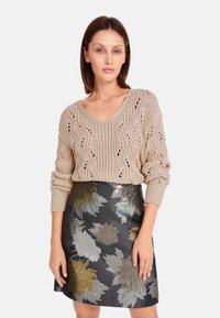 Beige gestrickter Pullover mit V-Ausschnitt und lässigen Ärmeln, kombiniert mit einem schwarzen Rock mit Blumenmuster und silbernen sowie goldenen Akzenten.