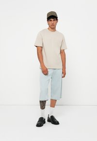 Beige cotton t-shirt, relaxed fit, paired with light blue denim shorts. Black shoes and plaid cap complete the outfit. White socks visible.