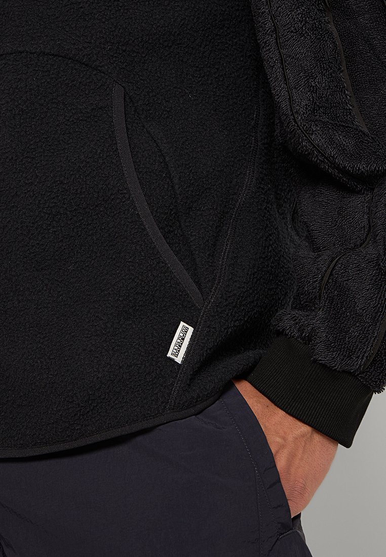 Poche d'une veste en polaire noire avec une étiquette de marque visible, poignet côtelé sur la manche, et main partiellement insérée dans la poche d'un pantalon sombre.