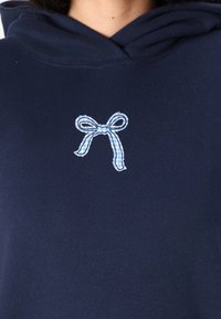 Marinblå hoodie med en mjuk textur har en blå och vit rutmönstrad broderad rosett på bröstet, vilket lägger till ett kontrasterande designelement.