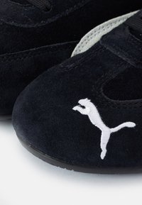 Gros plan d’une basket Puma en daim noir avec logo Puma blanc brodé sur l’orteil et semelle en caoutchouc visible sur une surface blanche.