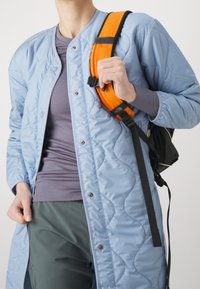 Lång, ljusblå vadderad kappa med tryckknappar, över en lila skjorta, kombinerad med mörkgröna byxor och en orange ryggsäck med rem.