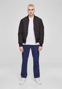Chaqueta bomber negra con cierre frontal, puños de canalé y bolsillo lateral, combinada con una camisa blanca y jeans azules. El modelo lleva zapatillas blancas.