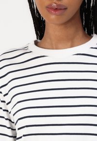 Una donna che indossa una camicia bianca con strisce nere orizzontali, mostrata dalla mascella fino alla parte superiore del petto, con i capelli intrecciati visibili.