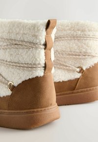 Brune faux suede støvler med en fluffy creamfarvet foring, der har flettede sidelister og guldtonet hardware.