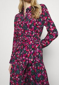 Frau in einem langärmeligen schwarzen Kleid mit floralem Druck in Rot, Pink und Lila, und einem passenden Stoffgürtel, der in der Taille gebunden ist.