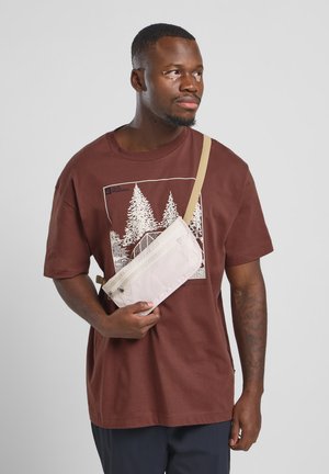 Crossbody tas in lichtbeige nylon met een ritssluiting en een verstelbare strap, gehouden tegen een bruine graphic tee met bomen.