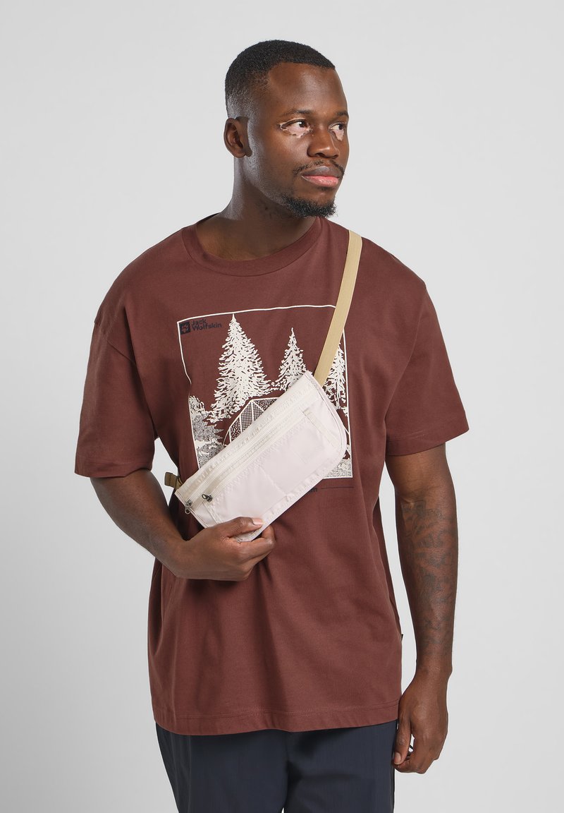 Crossbody tas in lichtbeige nylon met een ritssluiting en een verstelbare strap, gehouden tegen een bruine graphic tee met bomen.