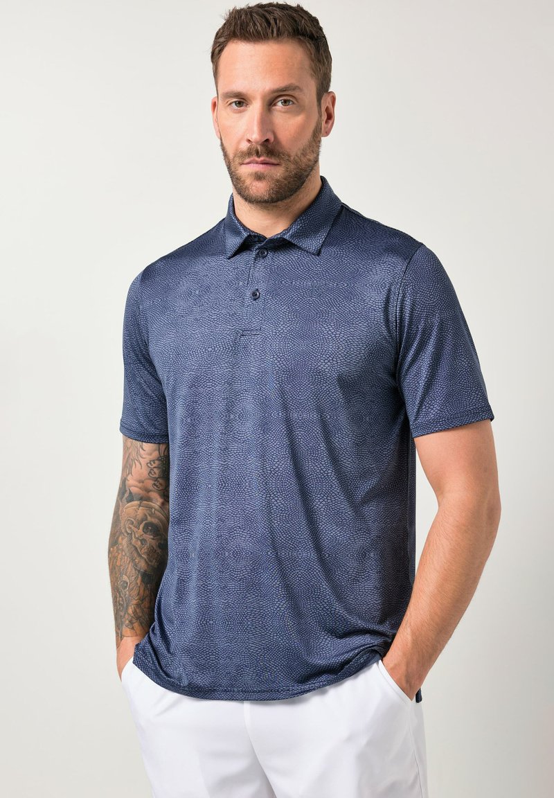 Mann mit kurzem braunem Haar und Bart, der ein blau gemustertes Polohemd und weiße Hosen trägt, mit den Händen in den Taschen vor einem einfarbigen Hintergrund steht.