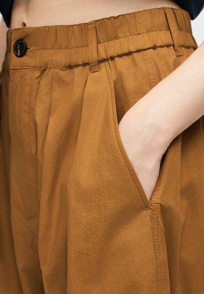 Χέρι στην τσέπη χαλαρών, ψηλόμεσων καφέ παντελονιών με κουμπί και θηλιές για ζώνη, φορεμένα με μαύρο τοπ που είναι μέσα στην μέση.